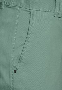 Pantalons en coton vert clair avec un tissu côtelé texturé, dotés d'une poche latérale, et accentués par un bouton noir près de la taille.