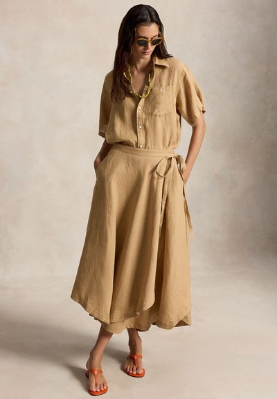 Moteris dėvi smėlio spalvos trumpomis rankovėmis marškinius, derančią sijono užrišamą dalį, geltoną karoliukų vėrinį, akinius nuo saulės ir oranžines basutes, stovinti su rankomis kišenėse.