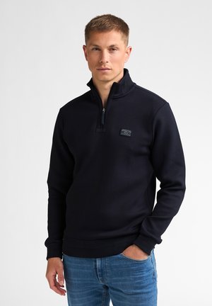Young man with short hair wearing a black quarter-zip pullover and blue jeans, standing with one hand in pocket against white background.