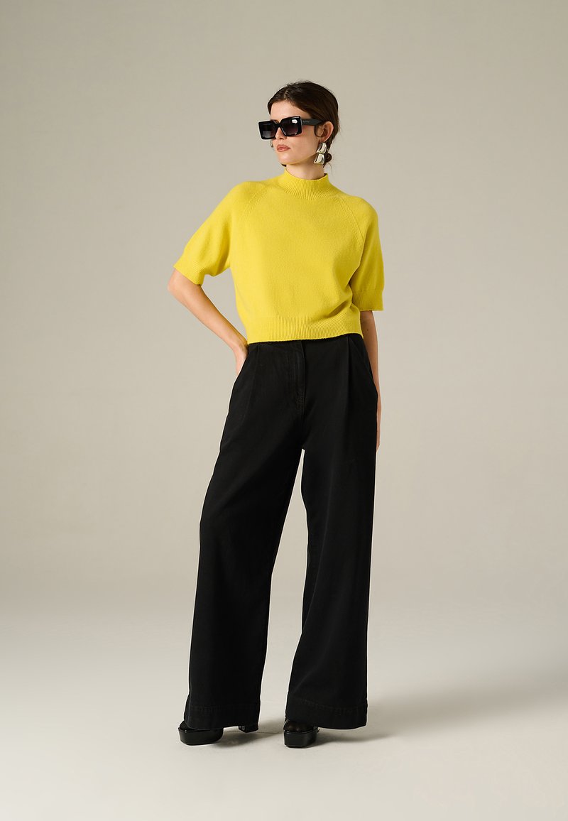 Maglione giallo a maniche corte in maglia, abbinato a pantaloni neri a gamba larga. Gli accessori includono grandi occhiali da sole neri e orecchini a cerchio in argento.