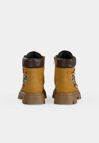 Paire de bottines en cuir jaune-brun avec semelles épaisses, cols rembourrés brun foncé et patchs brodés colorés sur les côtés.
