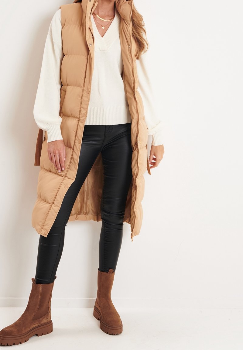 Khaki puffer vest with a high collar, zip closure, and side slits, layered over a white knit sweater and black faux leather leggings, worn with brown boots.