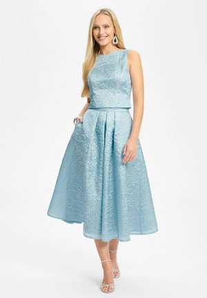 Femme aux longs cheveux blonds portant une robe jacquard florale bleu clair sans manches avec une jupe midi évasée et des talons ouverts argentés, souriante.