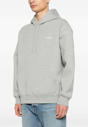 Mand iført lys grå Carhartt hættetrøje med forlomme og lyseblå jeans, stående mod en hvid baggrund.