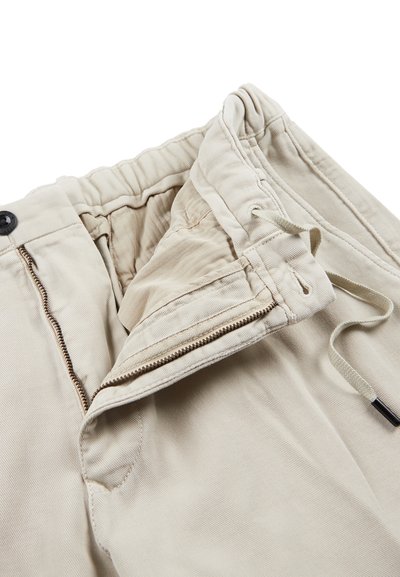 Beżowe spodnie z elastycznym pasem, suwakiem z przodu i sznurkiem do regulacji. Wykonane z miękkiej, teksturowanej tkaniny w wzór jodełki w okolicy pasa.