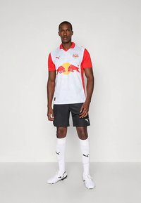 Muž oblečený v futbalovom drese Red Bull Salzburg s bielo-červeným dresom, čiernymi šortkami, bielymi ponožkami a futbalovými kopačkami, stojaci na jednofarebnom pozadí.