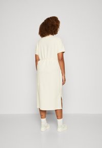 Vestido midi de crema clara con mangas cortas, cintura fruncida, aberturas laterales y textura suave, combinado con zapatillas blancas y tatuajes de tobillo visibles.