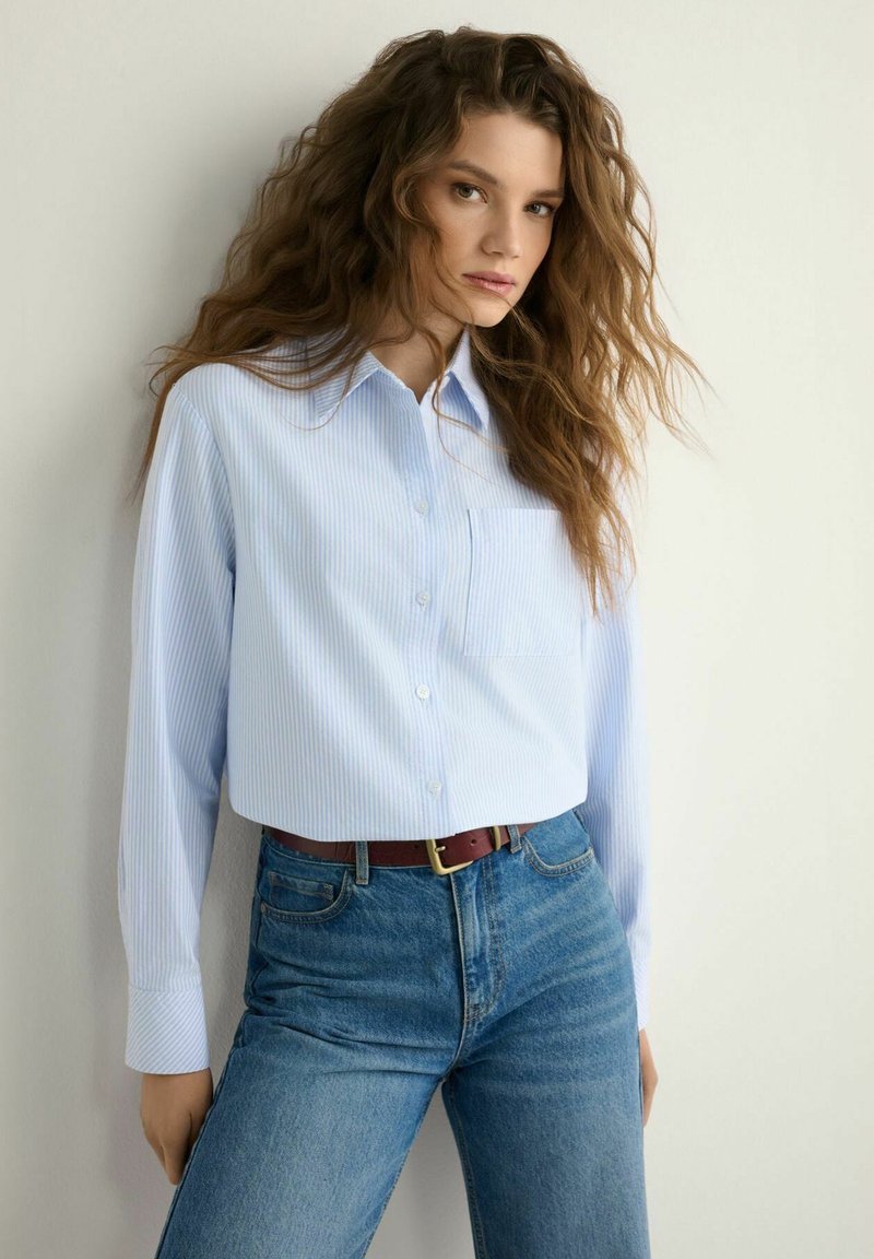 Kobieta o długich falowanych włosach, ubrana w jasnoniebieską koszulę na guziki, włożoną do wysokich dżinsów z brązowym paskiem, stoi na tle białej ściany.