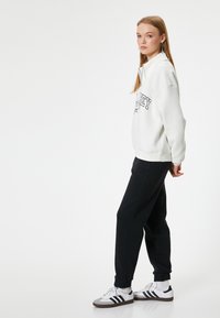 Weißer Sweatshirt mit schwarzem Schriftzug, schwarze Jogginghose und weiße Sneakers mit schwarzen Akzenten. Lässiger, lockerer Schnitt geeignet für den Alltag.