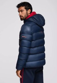 Uomo che indossa un giubbotto impermeabile blu navy con cappuccio e foderato di rosso, girato leggermente di lato, con capelli scuri e barba, su uno sfondo neutro.