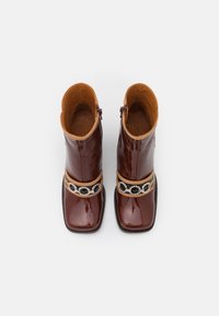 Bottines en cuir verni marron avec un bout carré, une bride en cuir marron clair et des pierres et perles noires décoratives le long de la bride.