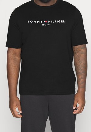 Czarna koszulka Tommy Hilfiger z logo i napisem "Est. 1985", noszona przez osobę z tatuażem na prawej ręce i ciemnymi spodniami.