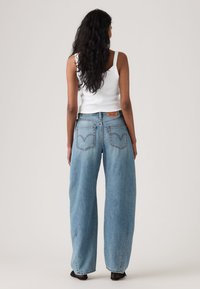 Γυναίκα με μακριά μαύρα μαλλιά φοράει λευκό κοντομάνικο τοπ και φαρδιά ανοιχτό μπλε τζιν παντελόνια, στέκεται αντικρίζοντας ένα απλό ανοιχτού χρώματος φόντο.