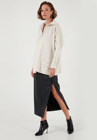 Pull léger beige, surdimensionné, avec un col zippé et des clous décoratifs sur les poches, associé à une jupe en cuir noire avec une fente sur le côté.