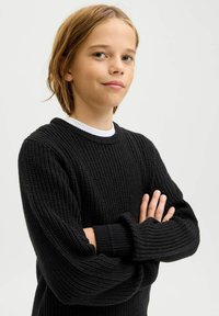 Schwarzer Strickpullover mit strukturiertem, geripptem Design. Runder Halsausschnitt und lange Ärmel mit umgeschlagenen Säumen. Über einem weißen Hemd getragen.