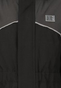 Veste noire avec des accent gris, dotée d'une fermeture éclair et d'une taille ajustable. Comprend une étiquette sur le côté supérieur gauche.