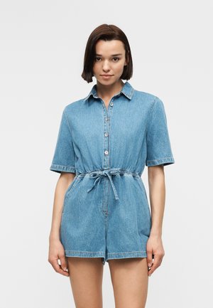 Junge Frau mit kurzem dunklem Haar, die einen blauen Jeans-Overall mit kurzen Ärmeln, vorderen Knöpfen und Taillengürtel trägt, vor einem schlichten Hintergrund stehend.