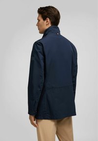 Marineblauwe lichtgewicht jas met een hoge kraag, een gladde textuur en een subtiel merklabel op de mouw; heeft een klassiek, minimalistisch ontwerp.