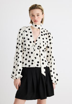 Fiatal nő fehér pöttyös blúzban, nyakkendővel és fekete pliszírozott szoknyában, sima fehér háttér előtt áll.