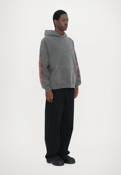 Sweatshirt à capuche gris avec une poche avant et des impressions graphiques rouges et jaunes sur les manches, associé à un pantalon large noir et des chaussures chunky.