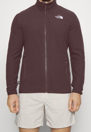 Osoba ubrana w ciemnoczerwony polar zapinany na zamek z logo The North Face na piersi oraz jasnobeżowe spodenki na jednolitym tle.