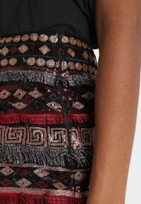 Zwarte stof met meerkleurige paillettenpatronen en franjes, met geometrische ontwerpen in rood, brons en grijs. Een ritsdetail aan de rand is zichtbaar.