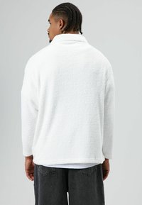 Witte, gestructureerde fleece trui met een hoge kraag en lange mouwen, gedragen over grijze broek. Heeft een losse pasvorm en zacht materiaal.