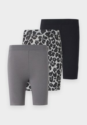 Joven con un top negro recortado, pantalones cortos de ciclismo con estampado de leopardo y chanclas negras, posando sobre un fondo blanco liso.