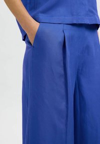 Mėlyni audinio kelnių su plačiomis kojomis dizainu, lygiu paviršiumi ir šoniniu kišenės akcentu. Medžiaga atrodo lengva ir pralaidi orui.