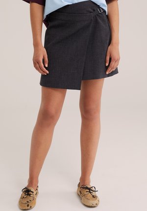 Donna che indossa una gonna a portafoglio grigio scuro con fibbia laterale e mocassini beige maculati, in piedi davanti a uno sfondo semplice.