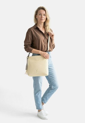 Kvinde med langt blondt hår iført en brun skjorte og lyseblå jeans, der holder en beige skuldertaske med tekstur og hvid rem.