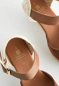 Sandali espadrilles in pelle marrone con tomaia intrecciata beige, fascia regolabile e soletta con texture. Presentano dettagli con logo dorato.