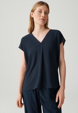 Frau trägt ein locker sitzendes, dunkelblaues V-Ausschnitt-Kurzarmshirt und passende Hose vor einem schlichten hellen Hintergrund.