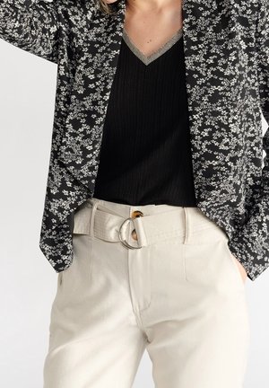 Vrouw draagt een zwarte blouse met bloemenpatroon over een zwart V-halsshirt, gestoken in beige broek met hoge taille en een stoffen riem.