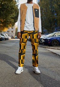 Beige fleeceväst med svart ficka, vit långärmad skjorta, mönstrade gula och svarta byxor, samt vita sneakers med grå detaljer.