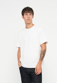 Camiseta de algodón blanca con cuello redondo y mangas cortas. Textura suave, diseño sin adornos. Modelo vistiendo jeans oscuros con un tatuaje detallado en el brazo.