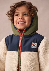 Detská bunda s námorníckym horným dielom, krémovým fleecovým telom a olivovo zelenou kapucňou. Obsahuje oranžový zips a nášivku s logom značky na hrudi.