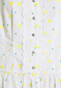 Blouse en tissu blanc avec un motif floral jaune, bordure en dentelle festonnée et boutons décoratifs. Comprend un ourlet froncé et des accents verts.