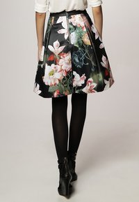 Jupe trapèze imprimée de fleurs avec une base noire, fabriquée en matériau brillant. Présente de grandes fleurs multicolores et un détail de fermeture éclair visible.