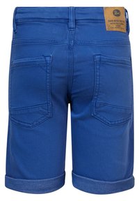 Shorts de mezclilla azul con dobladillo vuelto, dos bolsillos traseros y una etiqueta de marca de cuero marrón en la cinturilla. Textura suave, diseño ajustado.