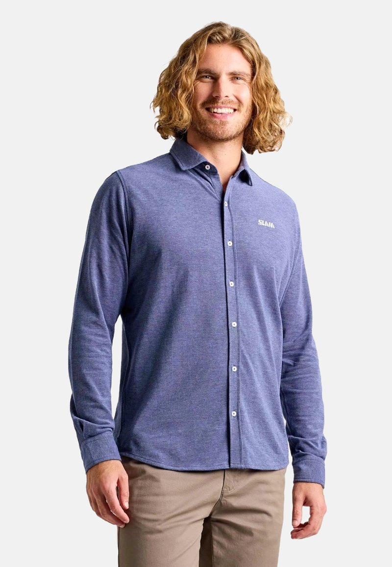 Chemise à manches longues, boutonnée en tissu bleu avec une texture subtile. Présente un col et un logo sur la poitrine. Associée à un pantalon en khaki.