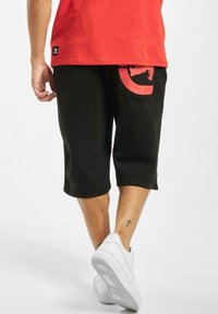 Pantaloni corti neri fino al ginocchio in cotone, con una grande grafica rossa sulla tasca posteriore. Abbinati a sneakers bianche.
