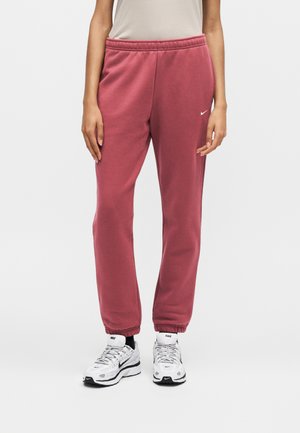 Person iført lyserøde Nike joggingbukser med elastiske manchetter og hvide Nike sneakers, stående mod en ensfarvet baggrund.