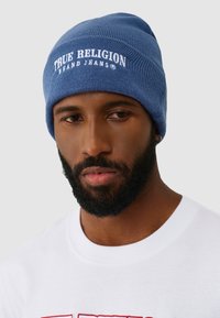 Man met baard die een blauwe muts draagt met de tekst "True Religion Brand Jeans" en een wit shirt met een rood ontwerp, tegen een lichtgrijze achtergrond.