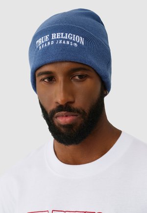 Man with beard wearing a blue beanie with "True Religion Brand Jeans" text and a white shirt with red design on a light gray background.