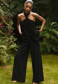 Crna halter top s drapiranim dizajnom u kombinaciji s širokim crnim hlačama. Oda dva komada su nježnog izgleda, smješteni u zelenoj vanjskoj pozadini.