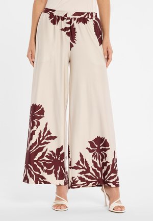 Beige Hosen mit weitem Bein und großen dunkelroten Blumenmustern an Taille, Oberschenkeln und Säumen, kombiniert mit weißen, geschnürten High-Heel-Sandalen.