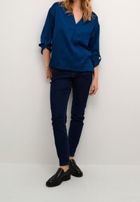 Blouse en satin bleu marine avec un décolleté en V et des manches retroussées, associée à un jean foncé ajusté et des mocassins noirs. Texture lisse, design élégant.