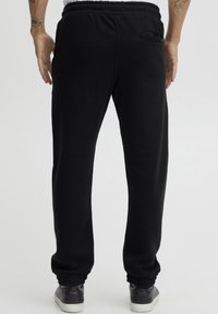 Pantalones de chándal negros con cintura y puños elásticos. Presentan un bolsillo trasero y una textura suave y lisa. Se llevan con zapatillas negras.