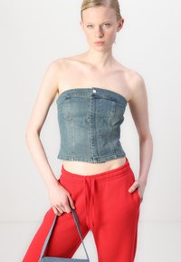 Denim crop top v svetlom modrom odtieni s priliehavým strihom, zapínaním na gombík a rovnými okrajmi. V kombinácii s červenými nohavicami so sťahovacou šnúrkou a modrou taškou.